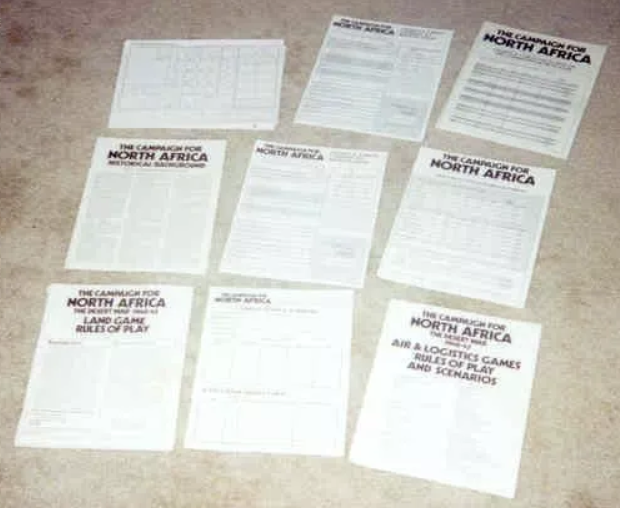 Six different rulebooks and three logistic-tracking sheets used in the game Campaign for North Africa. Very little can be read from the sheets at this distance other than the game’s name and titles for the rulebooks (e.g., “Land Game Rules of Play,” “Air & Logistics Games Rules of Play and Scenarios”).