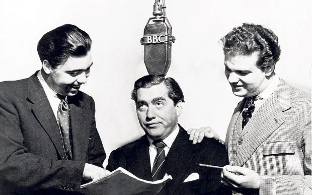 Alan Simpson, Tony Hancock and Ray Galton, wearing suits and stood around a BBC microphone. Simpson is holding and pointing at a script.