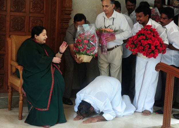 Jayalalithaa is seen seated as a man bows to her. Other men bear flowers and gifts in their hands to honor her