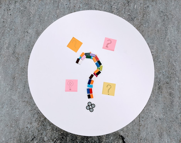 Photo of a question mark made of lego surrounded by four post-it notes with question marks on a round, white table.