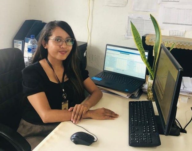 A woman sits at an office desk in front of two computer screens. Her hands rest on the desktop and she is looking straight at the camera.