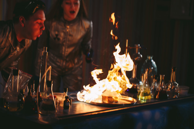 lab table with beakers and bottles around it, with large flames that a man and woman are attempting to blow out
