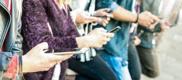 A group of 5 friends, all scrolling on their phones while standing against a wall together.