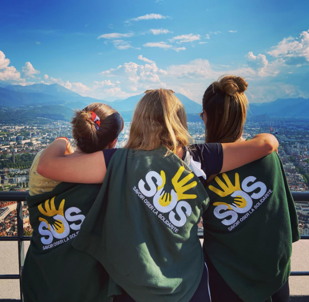 Three volunteers hugging in front of the mountains of Grenoble, with a sweater of the association.