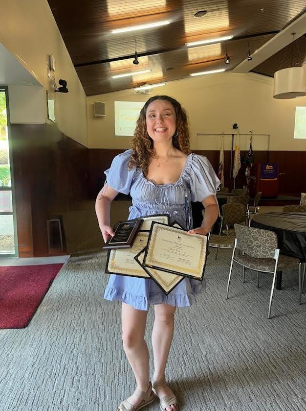 Emily Henderson stands in a blue dress, beaming widely as she holds up several awards she has received for her various leadership roles on campus.