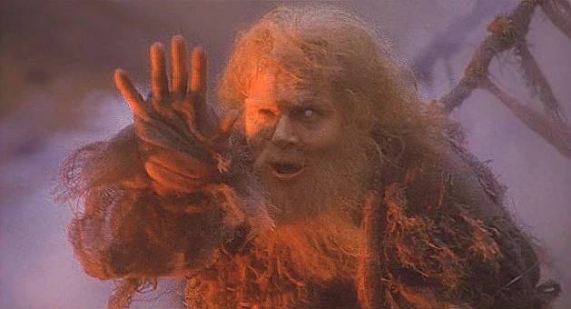 An ancient and imposing man holding his palm out to stop people crossing his bridge. It is a still from the movie Monty Python and The Holy Grail.