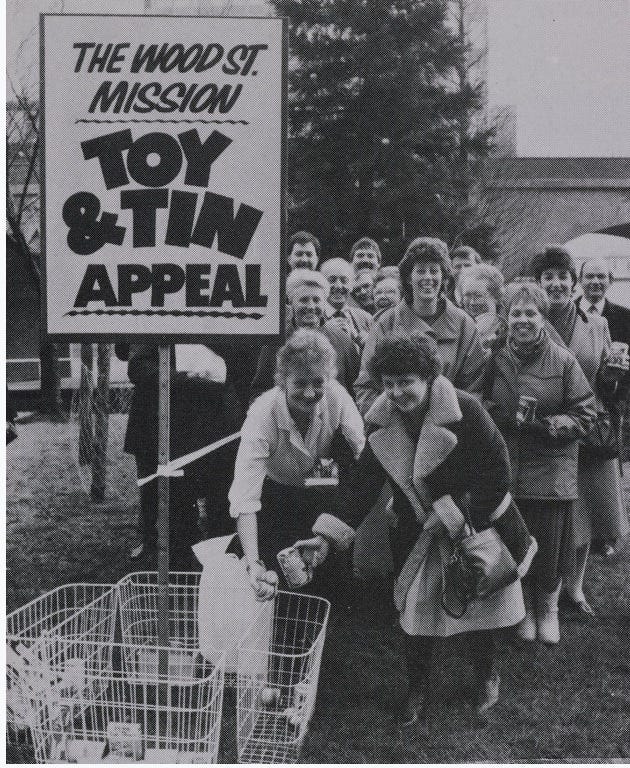 Group of mainly women with baskets of food and toys with sign say ‘Toy & Tin Appeal’