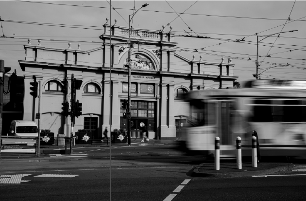 A greyscale photo of the Meat Hall at Vic Market with a moving tram in the foreground.