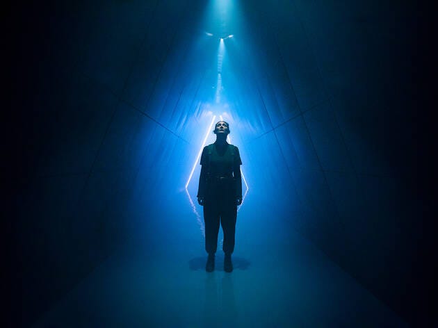 A woman stands in a darkened tunnel. The walls are angled so that the tunne is diamond-shaped. The space behind her is lit with neon blue light, so that she is framed and silouetted against it.