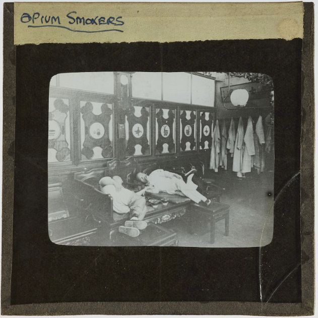 Black and white photograph of two men lying on a wooden bench inside a room. Slide is labelled ‘Opium Smokers’