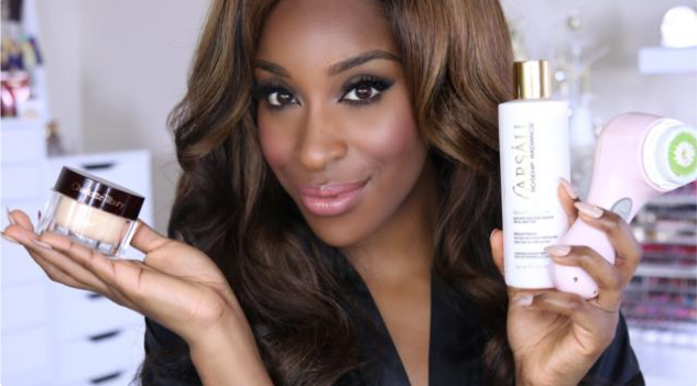 Black woman, Jackie Aina carrying facial products in her hands, one in her right, two in her left, with a drawer and makeup in the background
