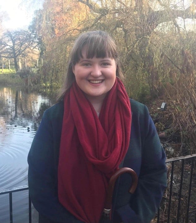 The author, smiling, wearing a red scarf, against the backdrop of a pond or stream and a thicket of trees.