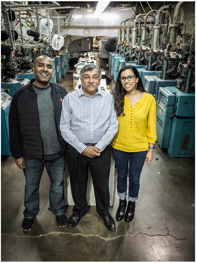 A high-angle portrait photograph of two men in plan clothing and one woman in a yellow cardigan. The man on the right in his late 30s, the man in the centre in his mid 50s and the woman on the right roughly 30. They are standing in a textiles factory floor.