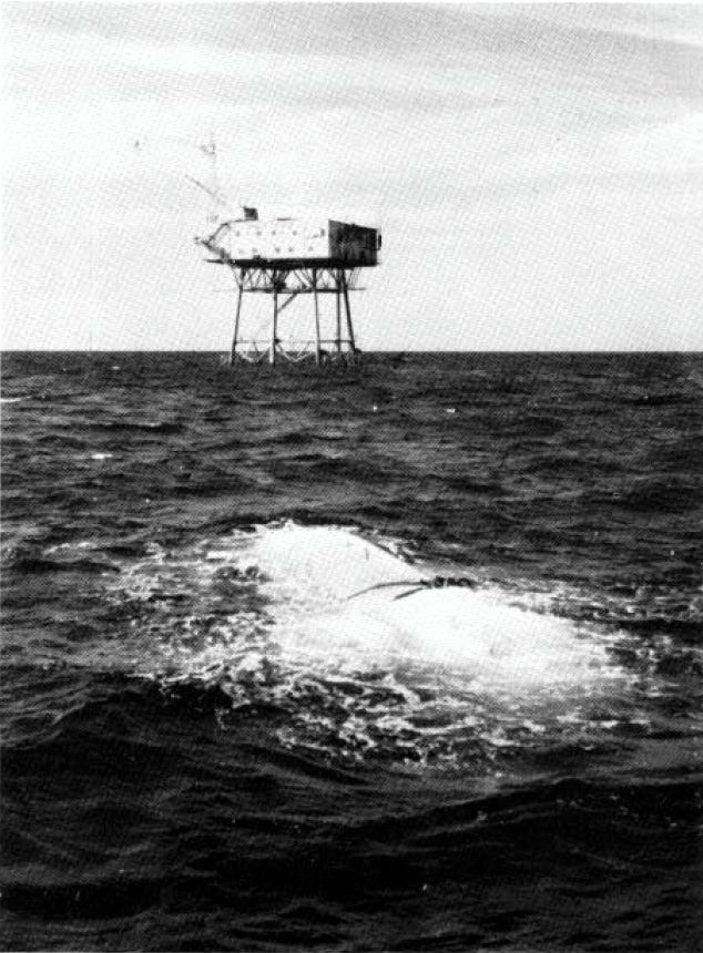 A grainy historic photograph shows a vessel submerging in water. A nearby tower is visible in the background.