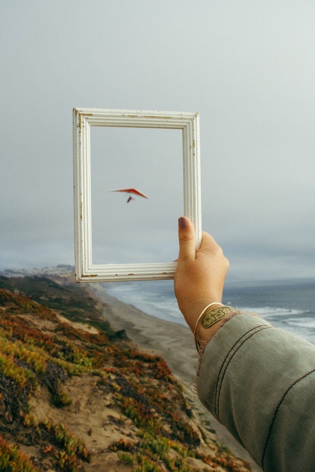 Uma mão segura uma moldura vazia que mostra uma asa delta pairar no céu em uma vista para praia ao fundo.