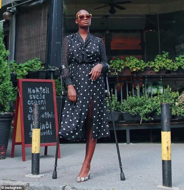 Mama Cax stands with shades on outside on a sidewalk, posed with her crutches and no prosthetic leg in a polka-dotted dress.