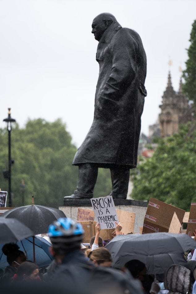 Picture of a BLM protest with banner in front of a statue saying ‘Racism is a pandemic too’