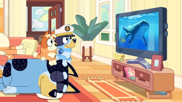 A still image from the cartoon show Bluey, showing the two children characters sitting on their dad’s head while watching a TV show