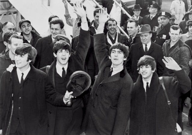 Black & white photograph of The Beatles arriving in America on 7 Feb 1964; from left to right, John Lennon looking left, Paul McCartney raising his left hand, George Harrison raising his right hand, Ringo Starr half-raising his left hand in front of a crowd of people