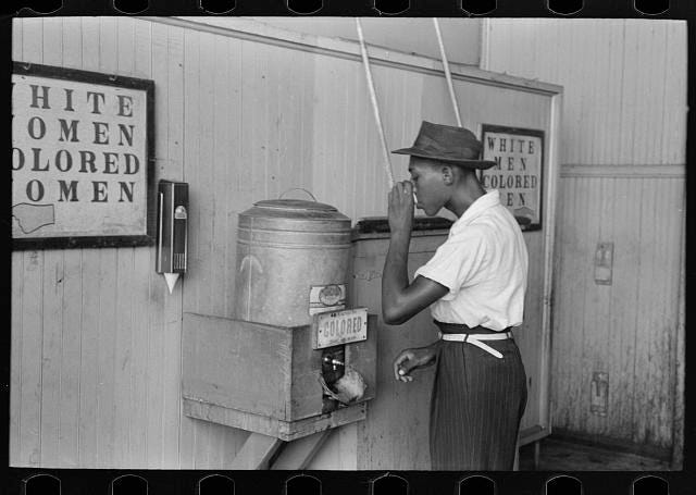 Black man drinking from a water fountain labeled ‘Coloured’ in between signs for “White women, colored women’ and “White men, colored men’