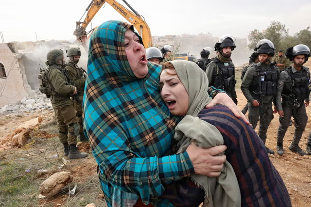 A imagem, no fundo, mostra um grupo de colonos israelenses vestindo uniformes militares. Na frente, há duas mulheres palestinas e muçulmanas, que parecem ser mãe e filha, lamentando aos prantos que sua casa está sendo demolida por colonos. O cenário parece ser ao ar livre sob o céu.