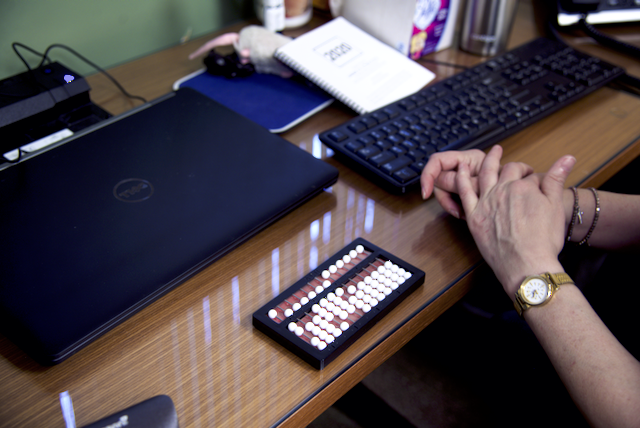 Chris sits with her hands folded on her desk, where a keyboard, laptop, and abacus also rest.