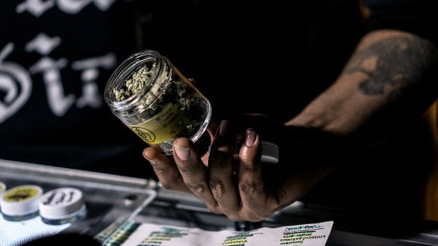 A man in a black shirt extending his arm with a jar of cannabis over a table.