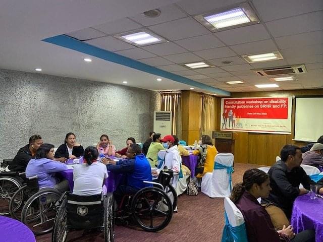 A photo of disability stakeholders seated at tables in a meeting room in Nepal. In the background a red banner with white English text reads “Consultation workshop on disability friendly guidelines on SRHR and FP.”