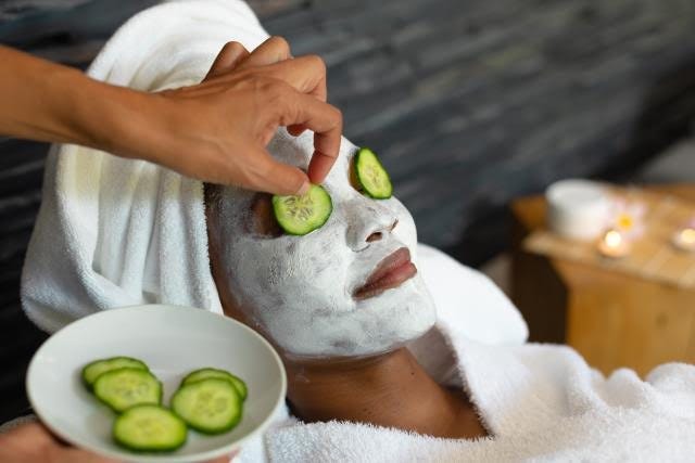 Stock photo of a woman in a white robe, white towel in her hair, white face mask, and cucumbers on her eyes. Another hand is holding the cucumbers on a plate, and placing one on the other woman’s eye.