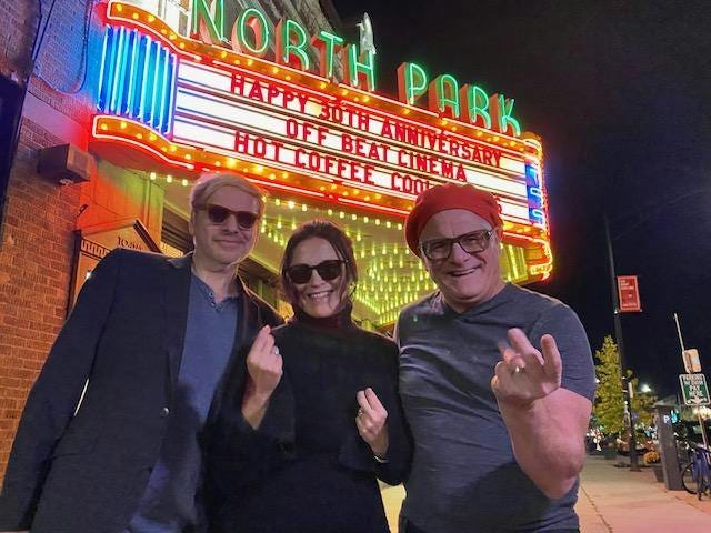 Three individuals stand before the illuminated North Park Cinema marquee at night. From left to right: a man with blond hair wearing a light jacket and dark pants, a woman in the middle with shoulder-length hair, donning a black turtleneck and sunglasses, and a man on the right wearing a red hat and glasses. The cinema’s sign is bright with neon lights, announcing ‘Happy 30th Anniversary’ and ‘Hot Coffee Cool Cinema’.