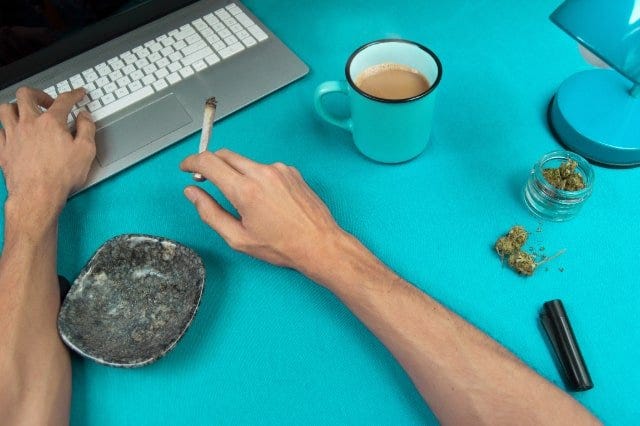 A man on a blue background holding a joint and using a computer next to a jar of cannabis, an ashtray, and a blue coffee cup.