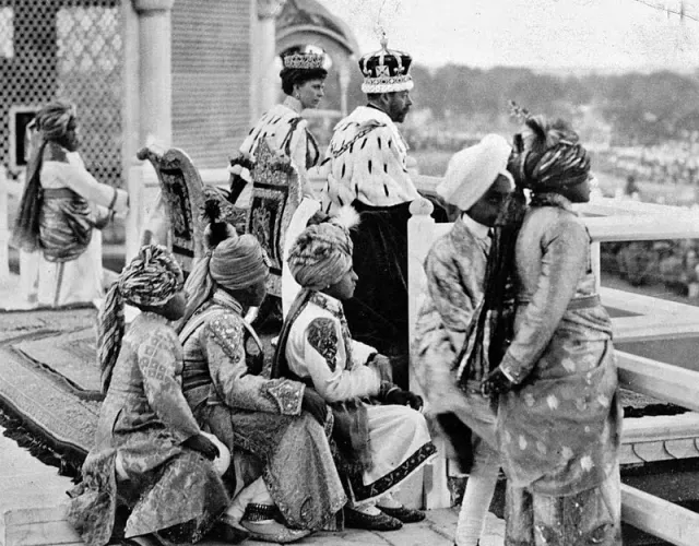 A historical black-and-white photograph showing British royalty, likely King George V and Queen Mary, dressed in regal attire with crowns and fur-trimmed robes, seated on ornate chairs. In the foreground, Indian nobility in elaborate traditional clothing and turbans observe an event, possibly the Delhi Durbar of 1911