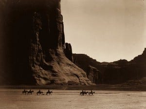Edward S. Curtis's photograph of Canyon De Chelly (1904) depicting horses and their cowboy riders across a southwestern landscape. Depicts a landscape commonplace in Cormac McCarthy's book, 'Blood Meridian."