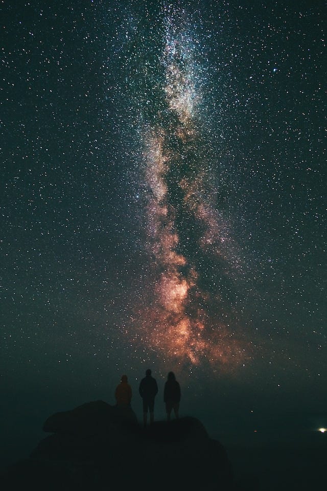 Unsplash color photo by Benjamin Davies, 2017, is a vertical composition with the colorful stars of the Milky Way reaching up into the night sky. At bottom center are three dark silhouetted figures standing on a hill, pondering the “inconceivably vast universe”. Title: three person looking stars and milky way. https://unsplash.com/photos/__U6tHlaapI