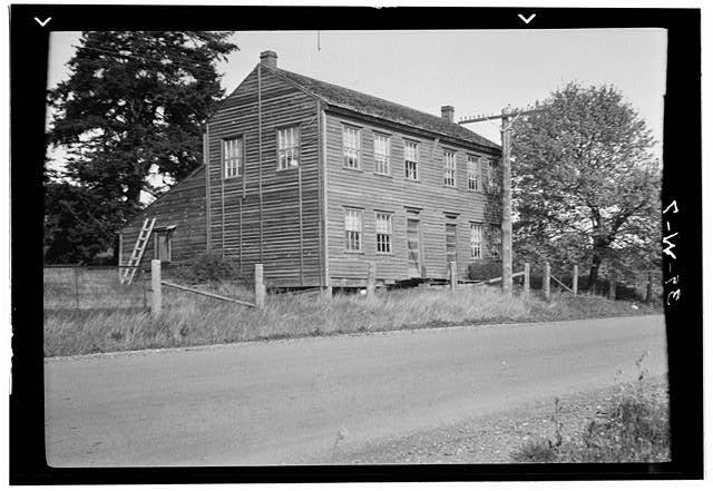A wood sided two story cabin-type building is shown in a black and white photograph. Windows adorn the top and bottom floors at the front of the cabin and adorn only the top floor on the side. A road and some fencing run in front of the cabin and a large power line stands in front as well. The cabin is surrounded by long grass and a few trees.
