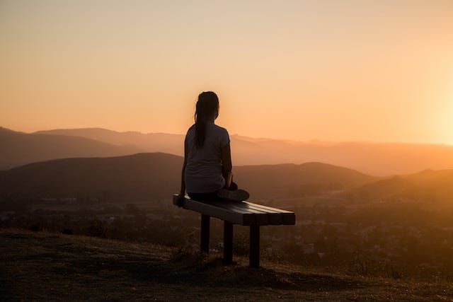 a woman sitting on a bench, looking into the distance. in the background a sunset over mountains. the light is golden.