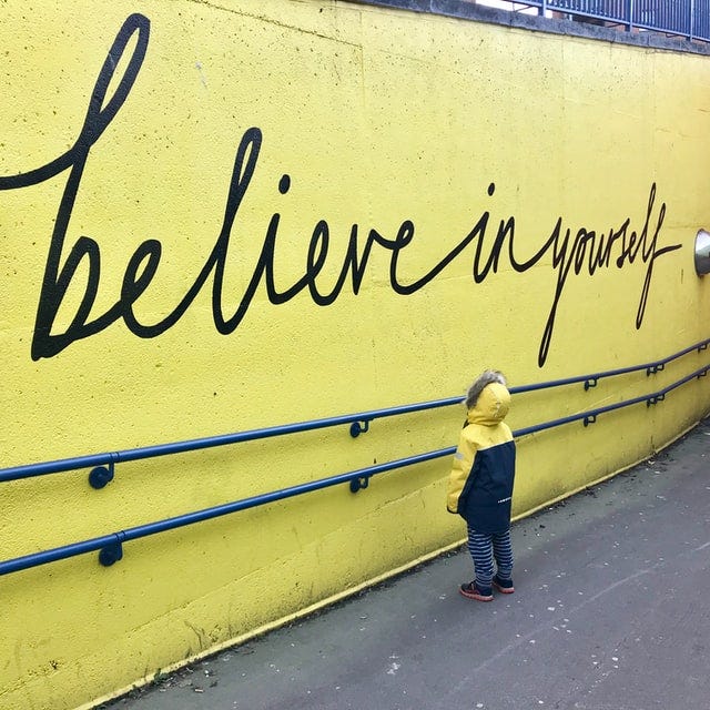 A child stands by a yellow wall. The words ‘believe in yourself’ are written in large script in graffiti.