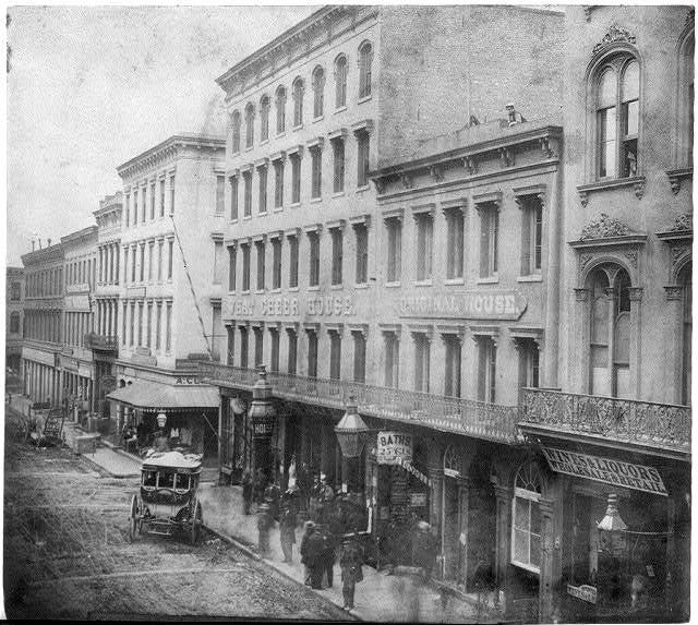 A city street made of dirt fronts two blocks of multi-floor city buildings in a black and white photograph. The largest building has lettering on its upper floors that reads “What Cheer House, Original House.” Signs on businesses at street level read “Baths” and “Wines & Liquors, Wholesale & Retail.” People are standing and walking on the sidewalk in front of the businesses and there is one carriage in front of the What Cheer House building.