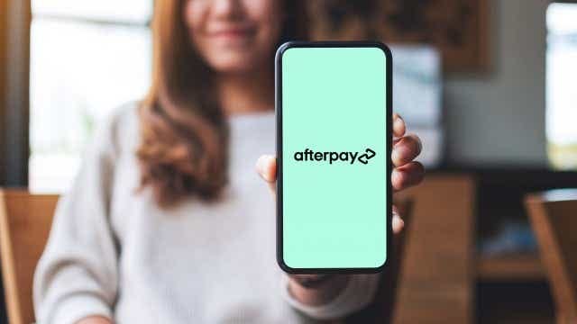 A person with long brown hair is holding up a phone to the camera. The phone has a mostly white screen with the Afterpay logo in the centre (all lowercase with two arrows at the end, one facing forwards, one facing backwards)
