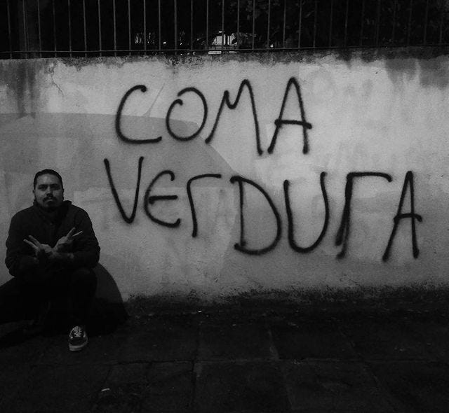 Eu na frente de um muro pixado com a frase “Coma verdura”