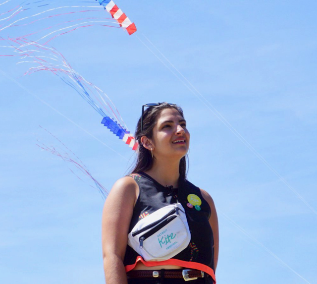 Margo Dalal, a young woman, looks to the sky with a smile during the Detroit Kite Festival. Kites fly in the background.