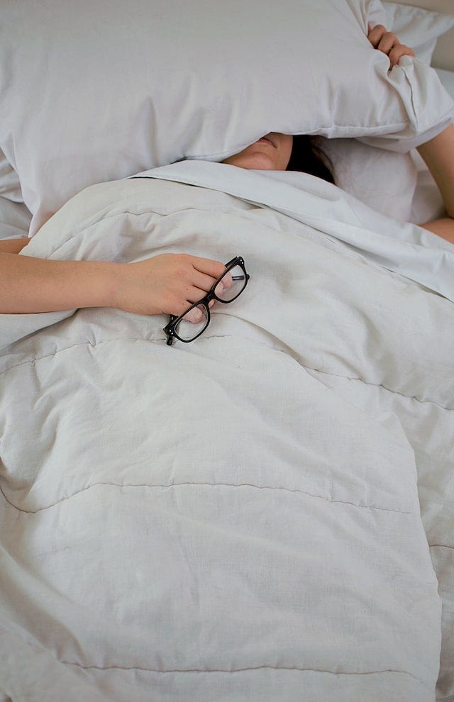 Woman under cream-colored comforter holding black rimmed glasses.