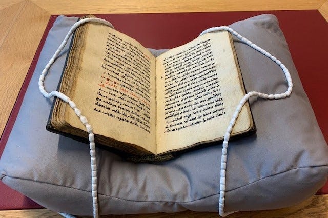 An open book supported on a book cushion, with snake weights positioned across the corners of left and right pages, allowing the book to safely stay open for study, without damaging the text