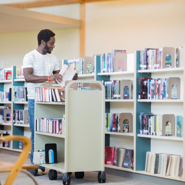 Full length of young male librarian with trolley arranging books in library