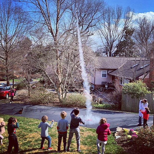 a toy rocket leaves a trail of smoke as it is fired off in the middle of a driveway, as children and adults look on with excitement.