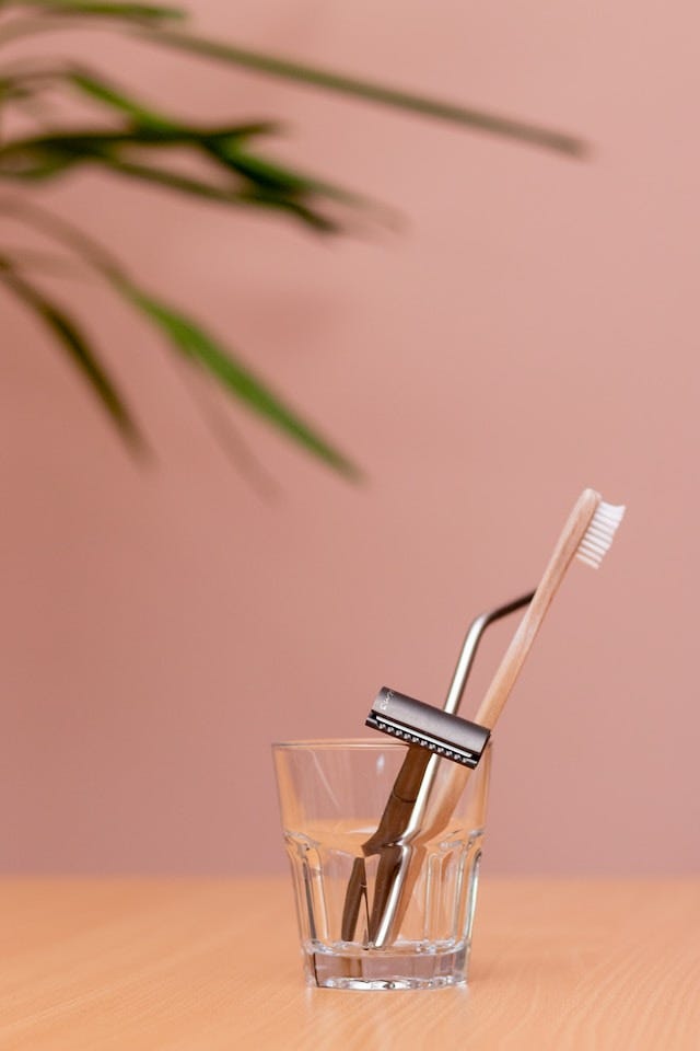 A clear glass cup holds a bamboo toothbrush, a metal safety razor, and a metal straw.