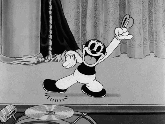 A screen capture from a Bosko cartoon, showing him performing on stage. He is a small, black, round person. He is wearing white kid gloves, has a large mouth ringed with big lips and has black skin.