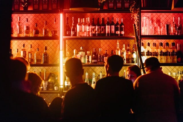 A group of men in a bar lit by neon lights, with their backs to the camera.