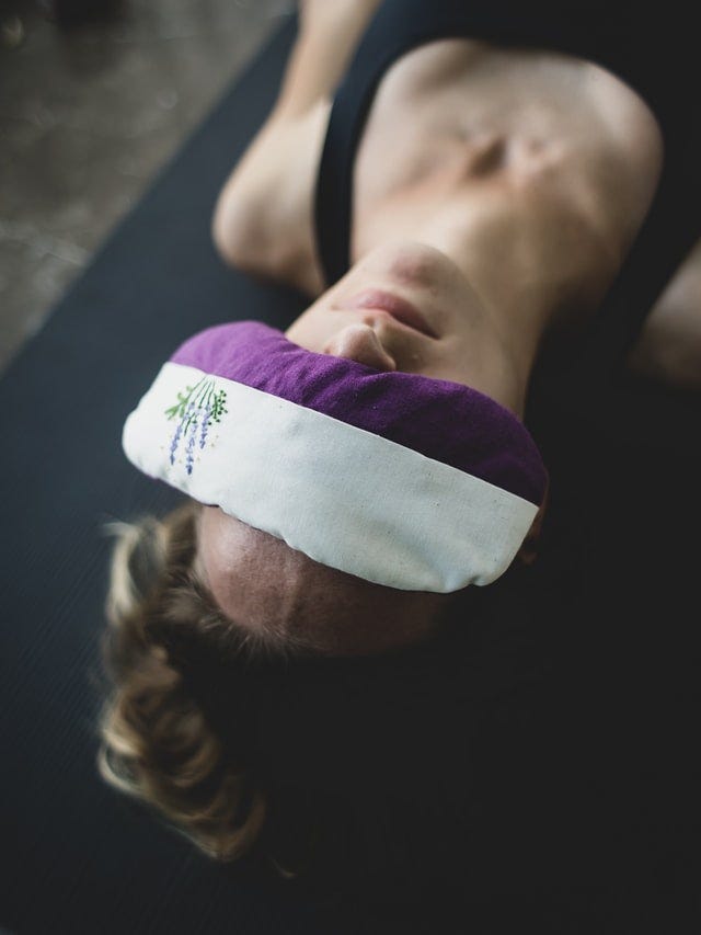 A woman, from the chest up, is seen lying on her back on what appears to be a yoga mat. Over her eyes there is a white and purple eye mask, with a small depiction of some lavender flowers stitched on one side.