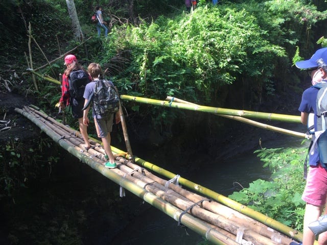 Children walk across a bridge made of 4 thick bamboo poles lashed together, drab green muddy river flows 15 feet beneath